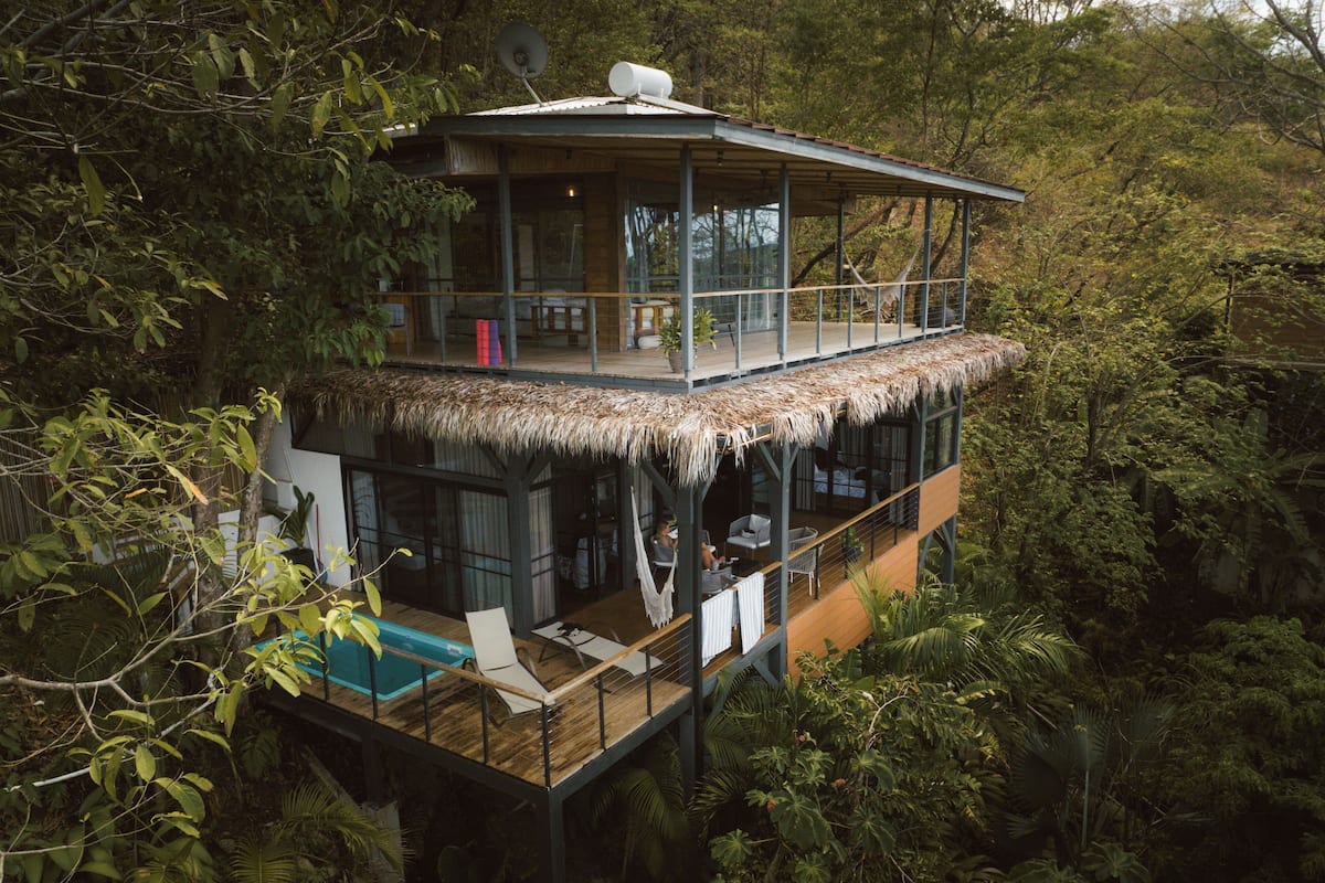 Uma casa na árvore de dois andares com telhado de palha, piscina e varanda com vista para uma área arborizada.
