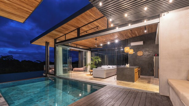 Ein moderner Poolbereich im Freien mit Holzdeck, Glaswänden und einer Sitzecke mit einer Couch und einem kleinen Tisch.