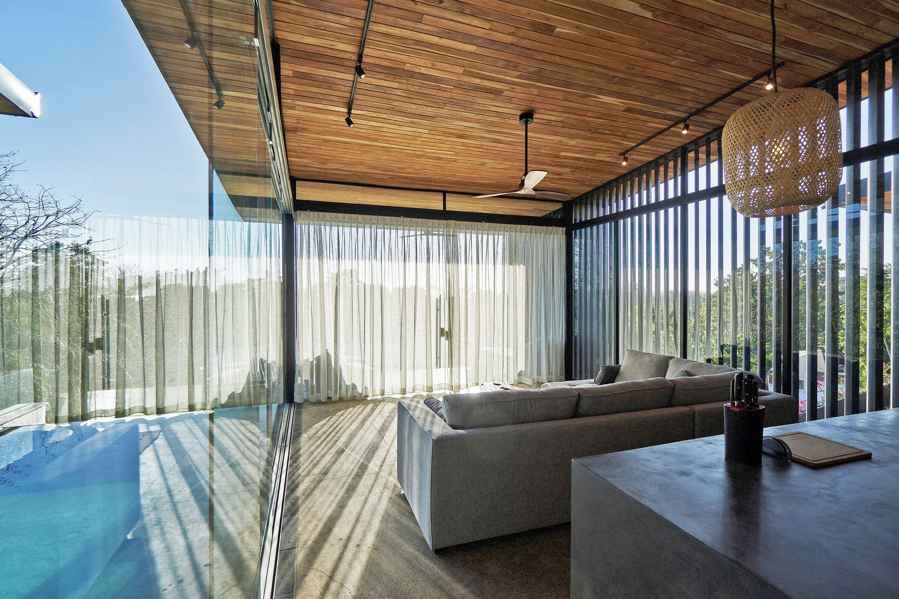 A modern living room with a large sectional sofa, a wooden ceiling, and large windows with sheer curtains.