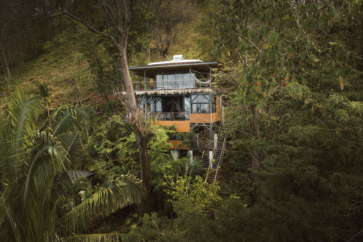 Uma casa de madeira de dois andares rodeada por vegetação tropical densa.
