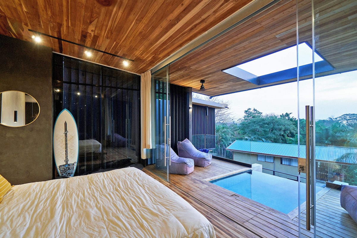 Quarto moderno com cama grande, parede de vidro e vista para uma piscina.