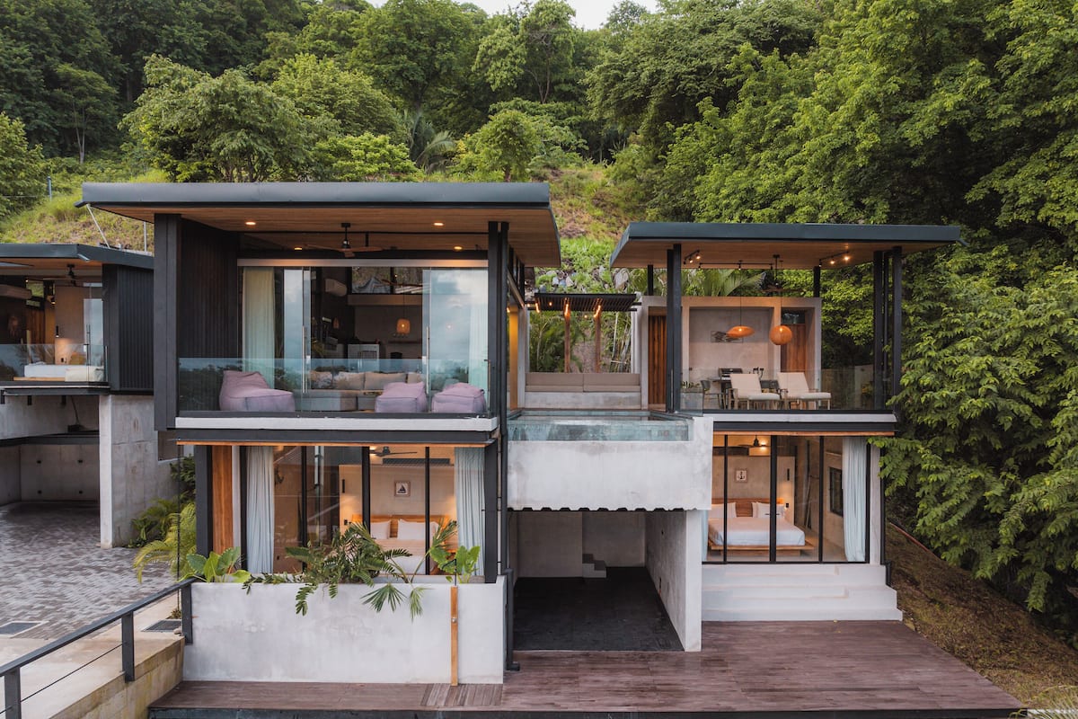 Uma casa moderna, com vários andares e varandas de vidro, rodeada por vegetação exuberante.