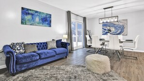 A modern living room with a blue sofa, a dining area with white chairs, and abstract paintings on the walls.
