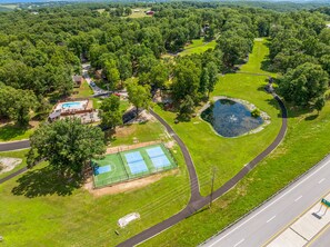An aerial view of a property with a swimming pool, tennis courts, a pond, and a road.