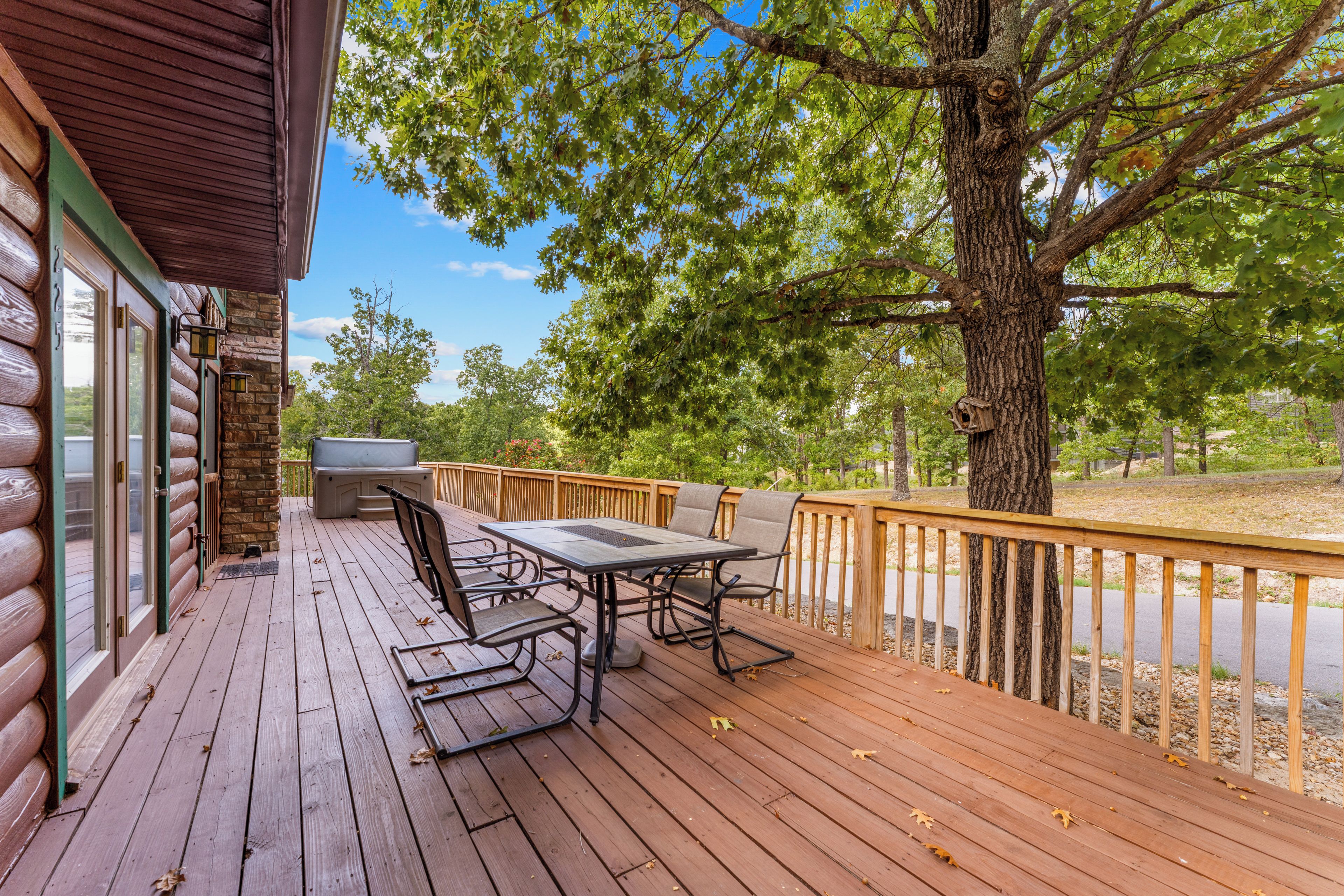 Una terraza de madera con una mesa y sillas, un jacuzzi y vistas a árboles y una carretera.