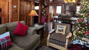 A rustic cabin interior with a high ceiling, wooden beams, a cozy seating area with a couch and chairs, a Christmas tree, and a kitchen area in the background. - Tailored Decor, Private Hot Tub, Authentic Log Cabin, Grill, Near Big Cedar&Thunder Ridge (Ridgedale)