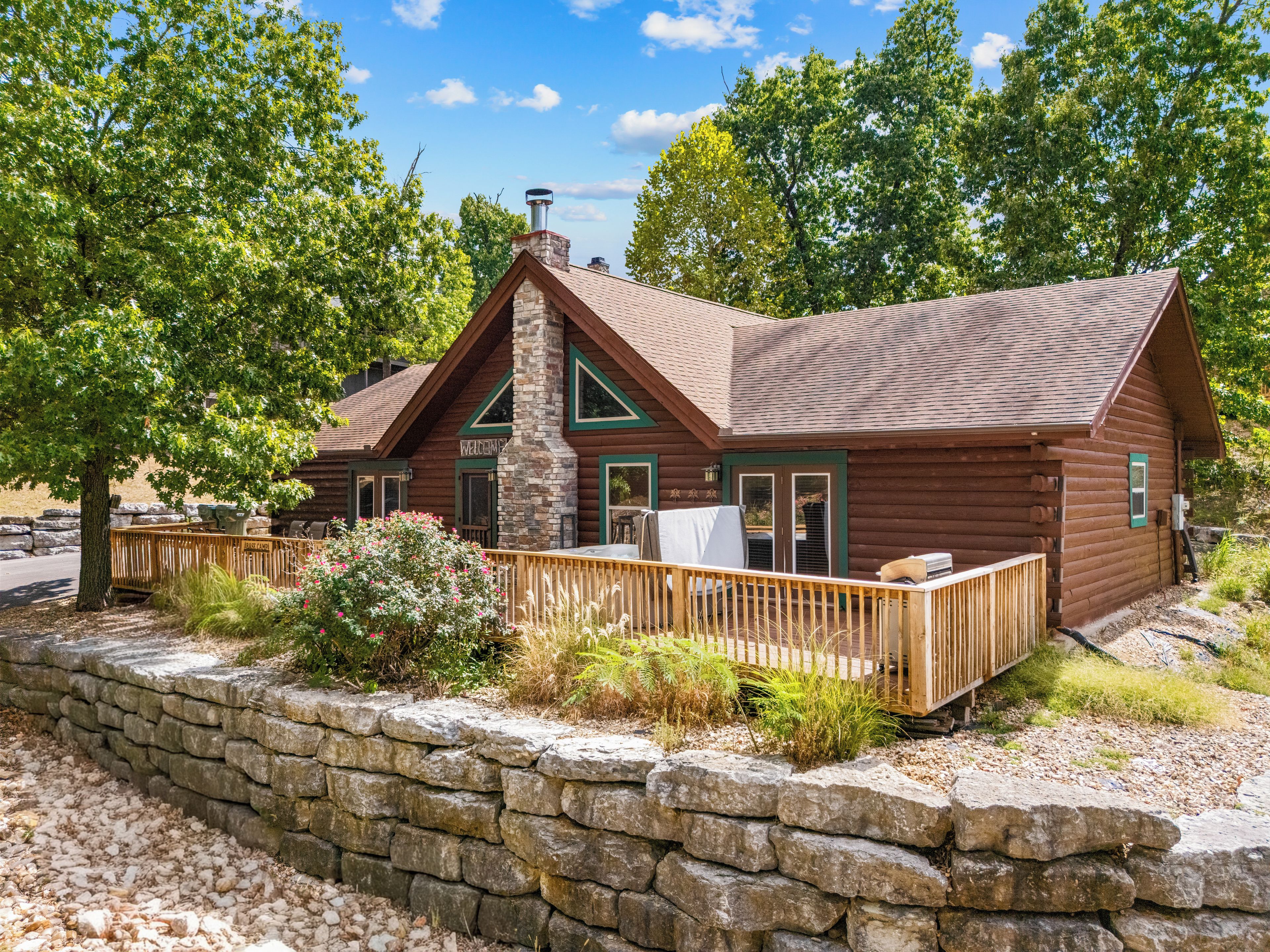 A log cabin with a stone retaining wall, a wooden deck, and a welcoming sign.