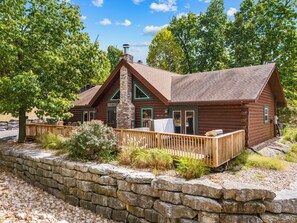 A log cabin with a stone retaining wall, a wooden deck, and a welcoming sign.