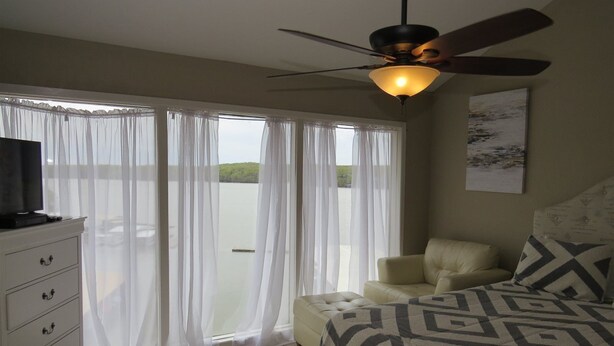 Un dormitorio con cama, ventilador de techo, televisor sobre una cómoda y vista al exterior a través de cortinas translúcidas.