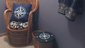 A wicker chair with a compass-themed pillow, a small round stool, and a wall-mounted towel rack with a dark towel. - Windjammer 33 (Osage Beach)