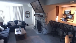 A living room with a stone fireplace, a sofa, a coffee table, and a kitchen area with a microwave and bar stools. - Windjammer 33 (Osage Beach)