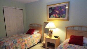 A room with two beds, each with a colorful bedspread and a red pillow. There is a nightstand with a lamp and a small table between the beds. A painting of fish and lily pads hangs on the wall above the nightstand. - Regatta BAY 404 (Lake Ozark)