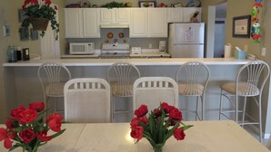 A compact kitchen with white cabinets, a microwave, a stove, and a refrigerator. There are four white chairs around a table with red flowers. - Regatta BAY 404 (Lake Ozark)