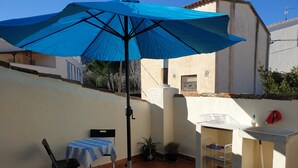 Eine Terrasse mit einem blauen Sonnenschirm, einem kleinen Tisch mit kariertem Tischtuch, einem Klappstuhl und einem Spülbecken mit einem Wäscheständer.