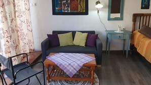 Ein gemütliches Zimmer mit einer Couch, einem kleinen Tisch mit kariertem Tischläufer, einem Sessel, einem Schreibtisch mit Lampe und einem Bett mit orangefarbener Decke.