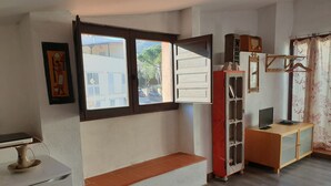 Ein Zimmer mit weißer Wand, einem Fenster mit Blick auf Gebäude, einer Holzbank, einem kleinen weißen Kühlschrank und einem Holzkabinett mit Spiegel und Fernseher.