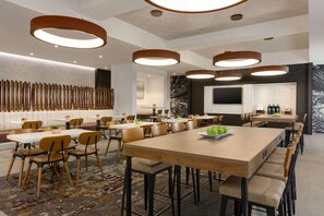 A modern dining area with wooden tables and chairs, a large central table with a fruit plate, and a decorative wall with vertical elements.