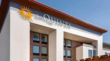 Fachada de hotel con el nombre "LAQUINTA" bien visible, que presenta un logo de sol y un diseño arquitectónico moderno con grandes ventanales y una pérgola de madera.