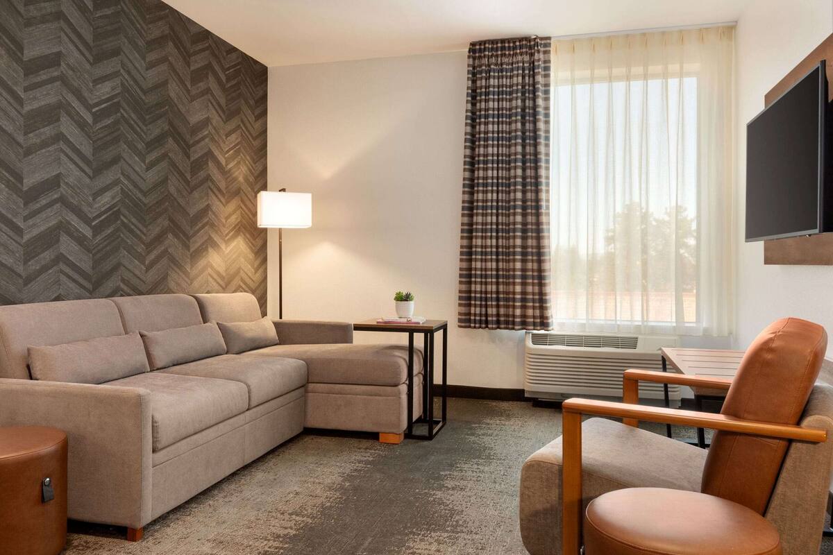 A modern hotel room with a sectional sofa, a single armchair, a small table with a lamp, a large window with curtains, and a flat-screen TV mounted on the wall.