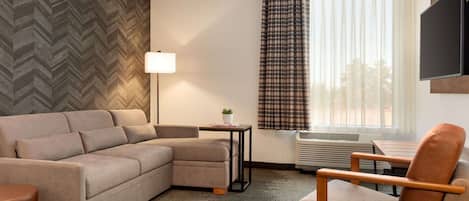 A modern hotel room with a sectional sofa, a single armchair, a small table with a lamp, a large window with curtains, and a flat-screen TV mounted on the wall.