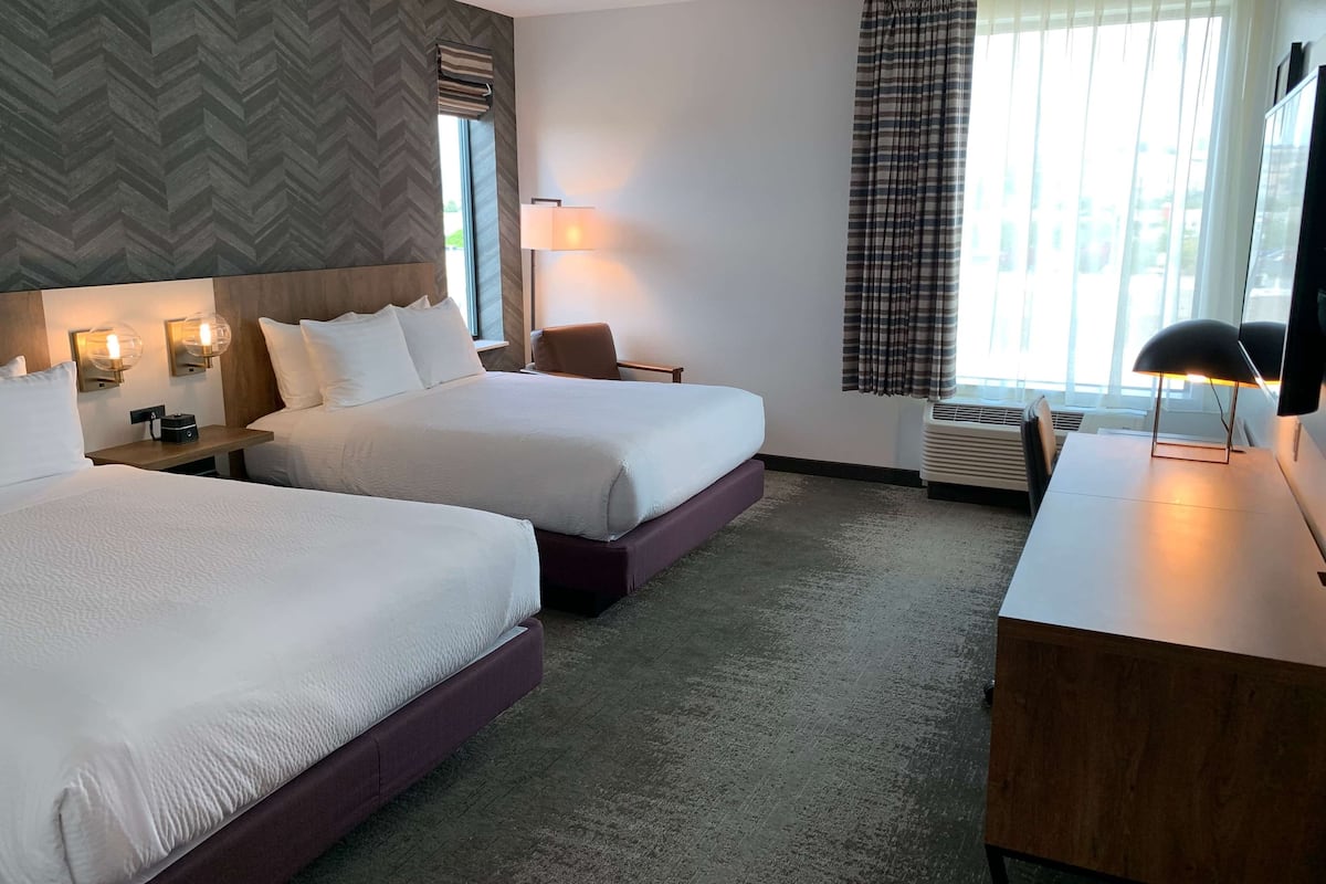 A hotel room with two beds, a desk, a chair, a lamp, and a window with curtains.