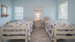 Um quarto com duas camas de madeira, uma mesa de cabeceira, um abajur fixado na parede e uma janela com persianas.