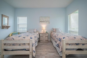 A bedroom with two wooden beds, a nightstand, a wall-mounted lamp, and a window with blinds. - Bay Front 4/3.5 home on Copano Bay! Pet Friendly (Rockport)