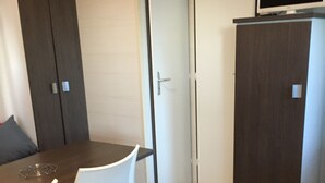 Una habitación pequeña con una mesa de madera, sillas blancas, un televisor pequeño en un mueble y dos puertas.