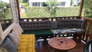 Una terraza de madera con una zona de estar acolchada que incluye un sofá amarillo y verde, una mesa redonda de madera y vistas a un seto y edificios al fondo.