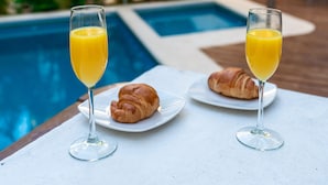 A table with two glasses of orange juice and a croissant, near a poolside with lush greenery and a white building in the background.