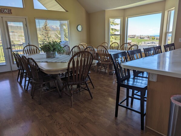 A spacious dining area with a large table and multiple chairs, a clock on the wall, and a view of the outdoors through large windows. - Golf Course Lakehouse with fantastic views of the lake, mountains & golf course. (Garden City)