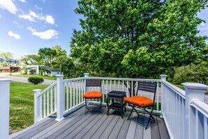 Una terraza de madera con barandillas blancas, dos sillas de mimbre con cojines naranja y vista a un árbol verde y una calle residencial.