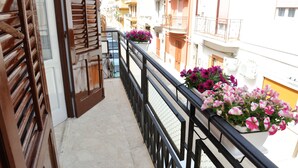 A narrow balcony with wooden shutters, a black metal railing, and flower boxes with pink and purple flowers.