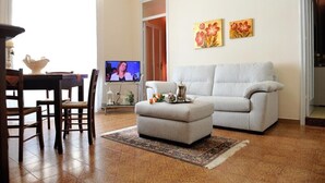 A living room with a grey sofa, a small ottoman, a dining table with chairs, and a TV. There are two floral paintings on the wall.