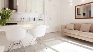 Une cuisine moderne avec des meubles blancs, un coin repas avec des chaises blanches et un canapé beige dans le salon.