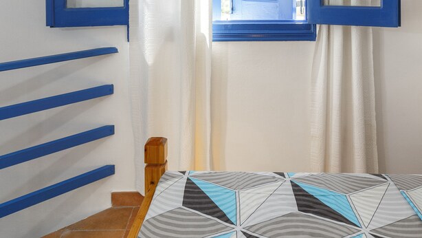 Una habitación con una cama, una ventana con contraventanas azules, cabecero de madera y cortina blanca.