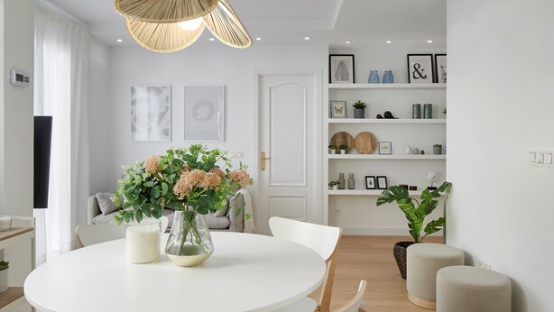 A modern dining area with a round white table, white chairs, and a vase with flowers. There is a white door in the background, a shelf with decorative items, and a hanging light fixture. - Moneo by FeelFree Rentals (San Sebastián)