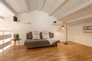 Une chambre de style loft moderne, avec un plancher en bois, un canapé-lit gris agrémenté de coussins blancs et noirs, une lampe sur pied et une petite plante posée sur une table d’appoint.