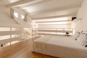 A bedroom with a large bed, wooden flooring, a white ceiling, and a staircase leading to an upper level.