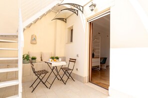 A small outdoor dining area with a table and two chairs, a white railing, and a door leading to an interior space.