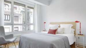 A modern bedroom with a large bed, two bedside tables, a chair, and a view of buildings through large windows. - Larroka by FeelFree Rentals (San Sebastián)