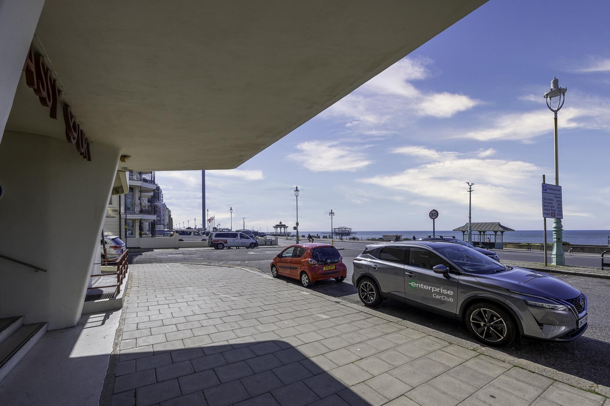 A parking area with cars, a building with a sign, and a view of the sea.