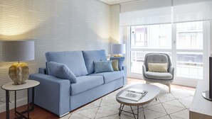 Uma sala de estar moderna com um sofá azul, uma poltrona cinza e uma mesa de centro redonda. Há duas luminárias sobre mesas laterais, e uma grande janela permite a entrada de luz natural.