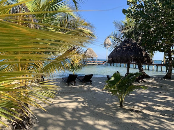 A beach with lounge chairs, palm trees, and a thatched structure over water.