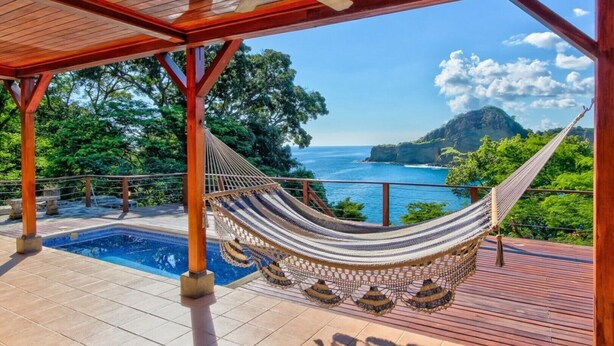 A hammock on a wooden deck with a pool and a view of the ocean and cliffs. - Oceanfront treehouse home w/ private pool, deck, near beach (Tola)