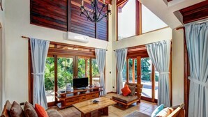 A spacious living room with a wooden coffee table, sofas, and a TV on a wooden stand. Large windows offer views of greenery outside. - Oceanfront treehouse home w/ private pool, deck, near beach (Tola)