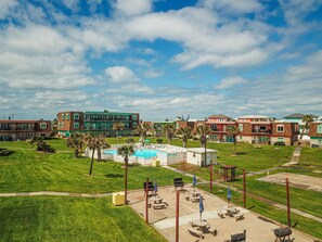 A pool area with lounge chairs and umbrellas, surrounded by green lawns and buildings with multiple stories. - Walk to Sandfest, Shared Pools, Boardwalk to Beach (port aransas)
