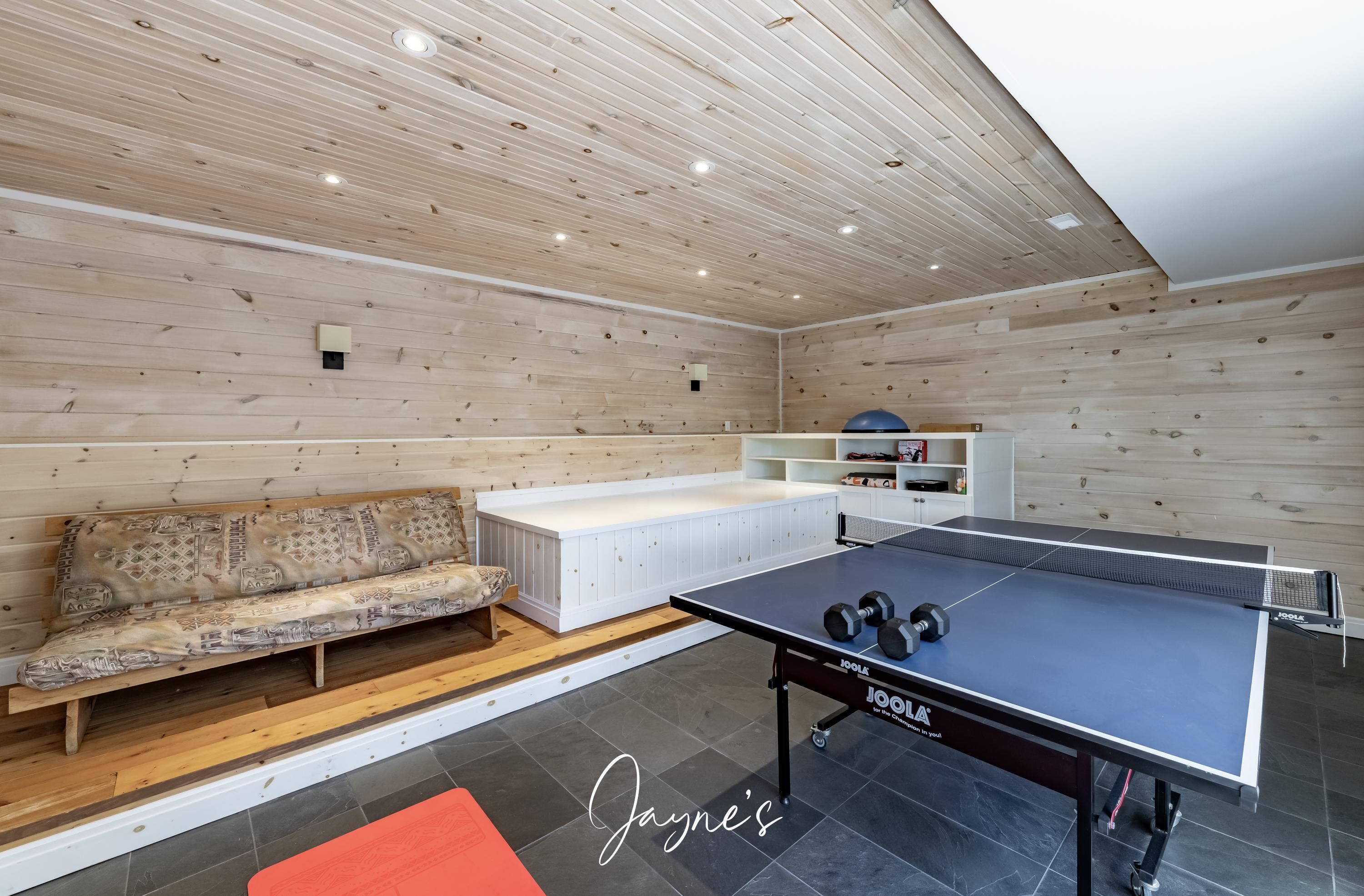 A ping pong table with a Joola brand table, a patterned sofa, and a white bench in a room with wooden walls and ceiling.