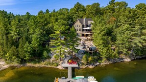 A lakeside property with a dock, boats, and a large house surrounded by dense forest. - Sensational Georgian Bay Cottage 6 Bdrms, 5 with Ensuites! Hot Tub! AC! (Parry Sound)