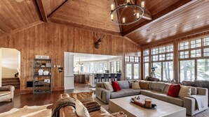 A spacious living room with a large sectional sofa, a coffee table, and a fireplace. There is a kitchen in the background with a large window. - Gorgeous 6 bedroom | 6 bathroom cottage with living quarters in Boathouse! (Seguin)